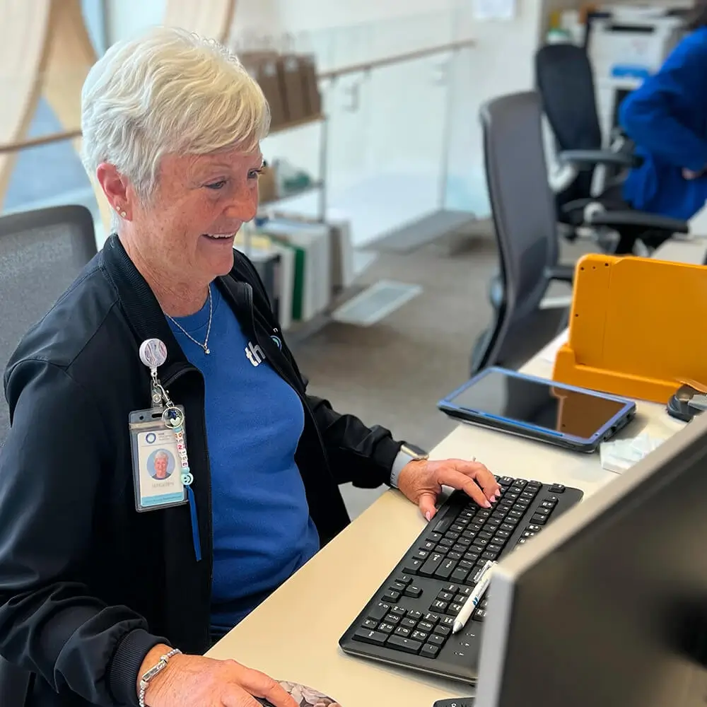 An older woman with short gray hair, wearing a badge and blue shirt, smiles while working at a computer desk in an office setting.