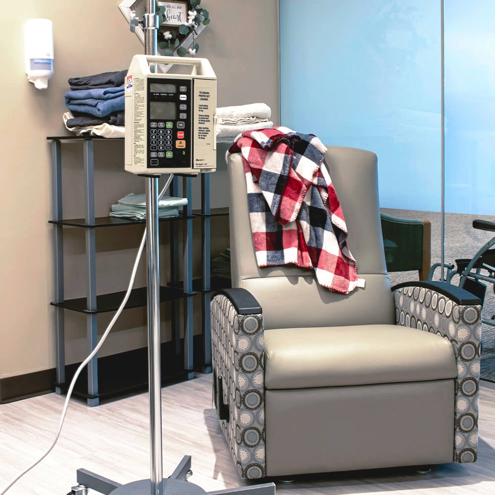 A medical infusion chair sits beside an IV stand in a clinical room. A red, white, and blue plaid blanket is draped over the chair, with folded towels on a shelf in the background.