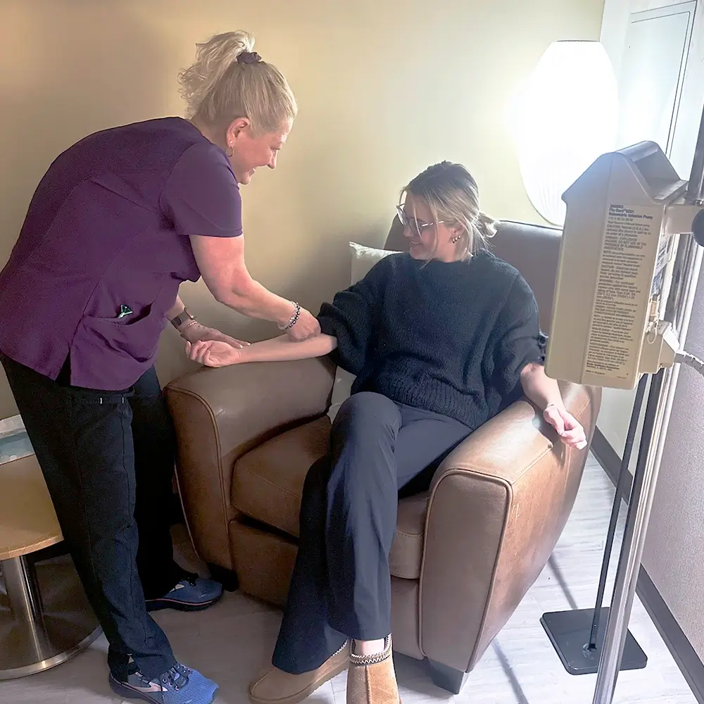 A healthcare worker in purple scrubs prepares to draw blood from a smiling woman seated in a brown armchair, with medical equipment and a lamp nearby in a clinic room.