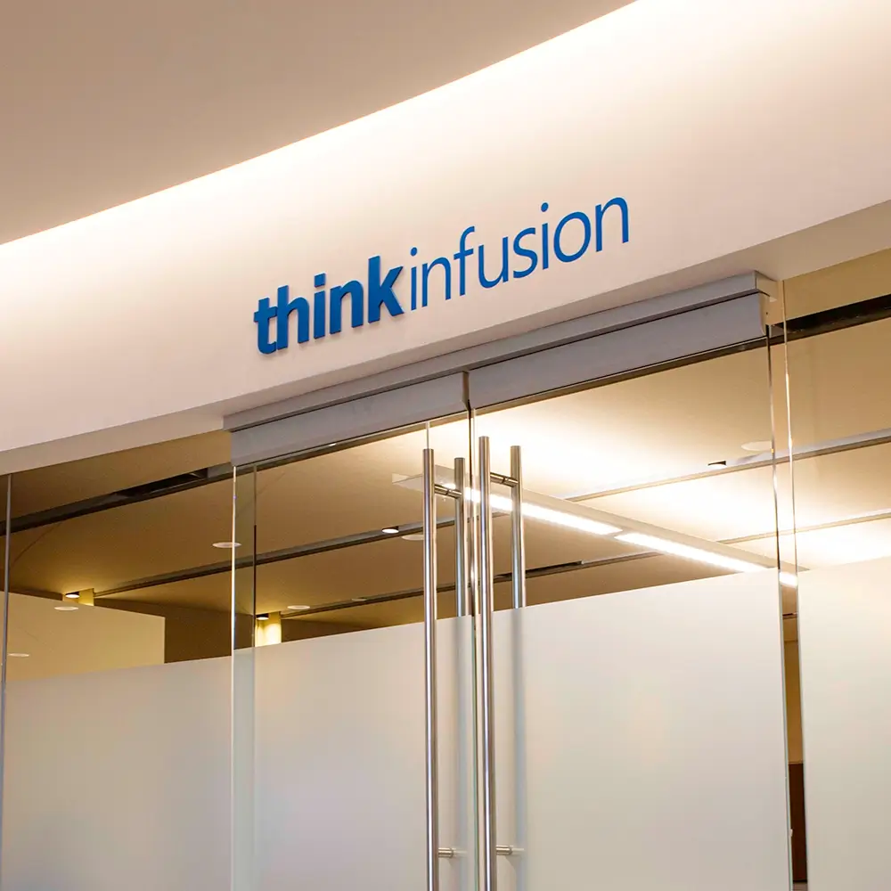 A modern office entrance with glass doors and a frosted glass panel. Above the doors, a sign reads “thinkinfusion” in blue letters on a white wall. Warm ceiling lights illuminate the space.
