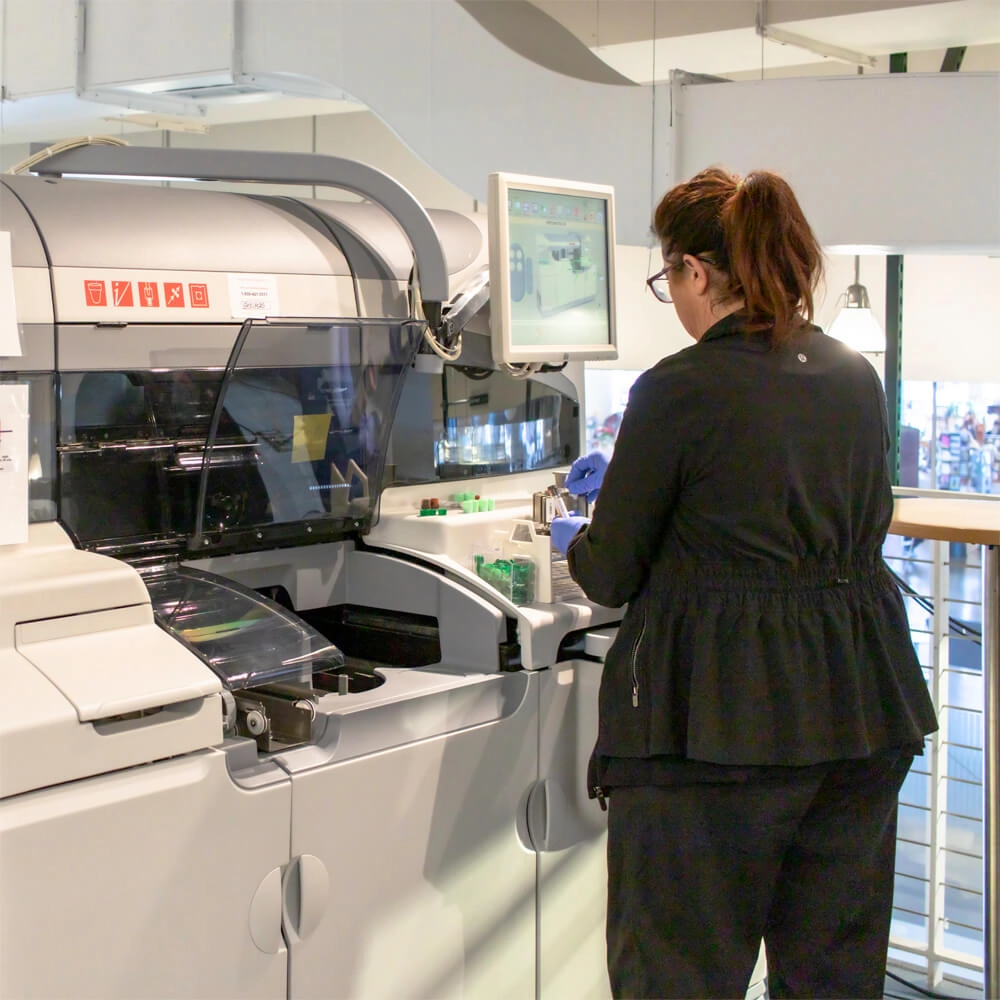 A woman in black clothing operates a laboratory analyzer machine, placing test tubes inside while looking at a monitor displaying data. The setting appears to be a modern laboratory.