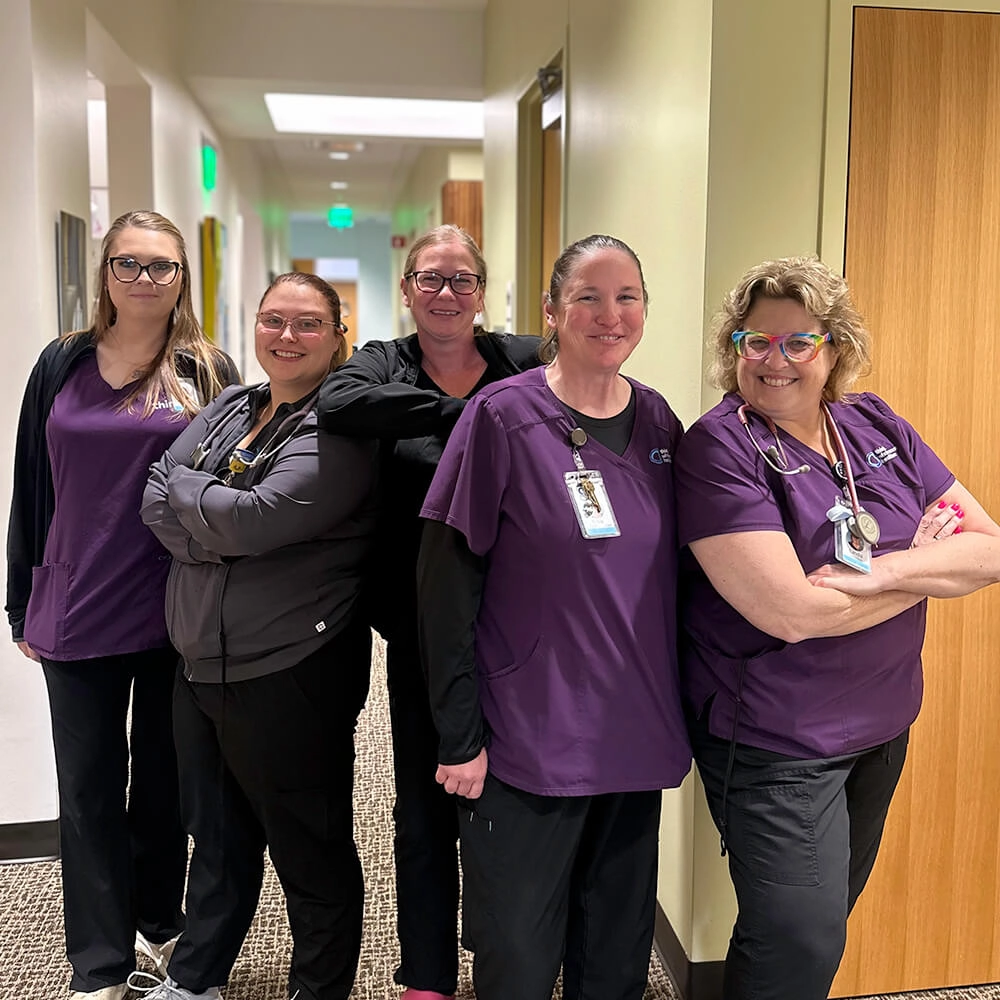 Five women in matching purple and black scrubs stand smiling together in a hallway, two with arms crossed and others with hands by their sides, wearing ID badges and stethoscopes.