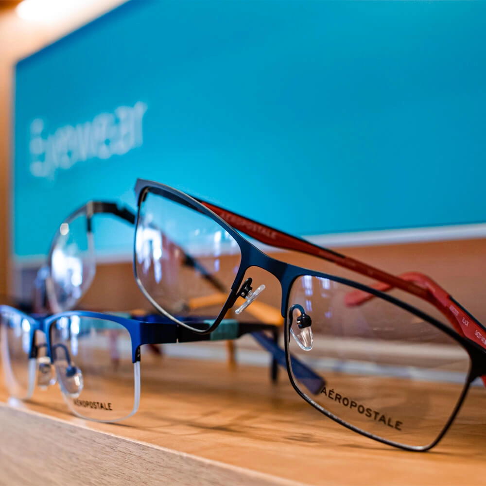 Close-up of three pairs of Aéropostale eyeglasses on a wooden shelf, with a blurred blue sign labeled eyewear in the background.