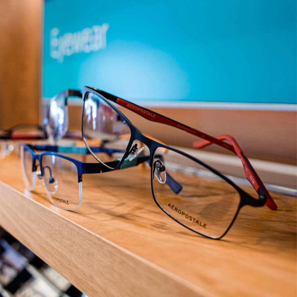Three pairs of Aeropostale eyeglasses are displayed on a wooden shelf in front of an aqua sign that says Eyewear. The glasses are in different styles and colors.