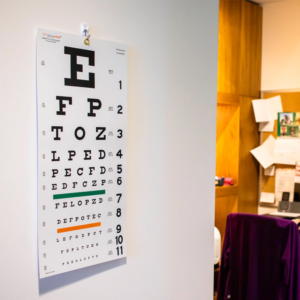 An eye chart with rows of letters decreasing in size is pinned to a white wall in an office, with a desk, papers, and a purple chair visible in the background.
