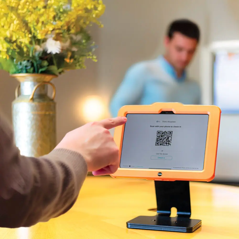 A person points at a tablet displaying a QR code on a stand, with another person blurred in the background and a vase of yellow flowers on the table.