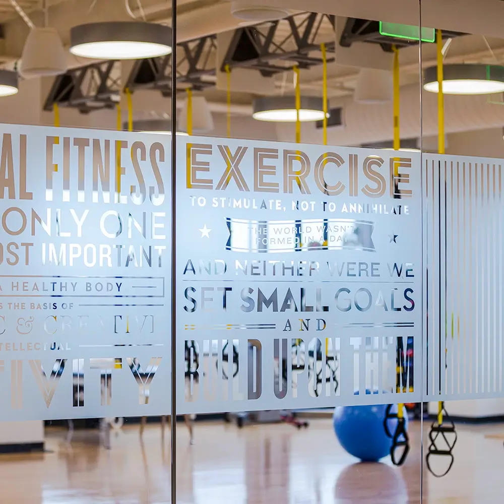 A reflection of a gym's motivational wall art, featuring phrases about fitness, exercise, and setting goals, on glass partitions.