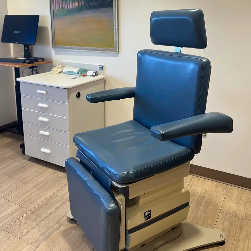 A blue padded medical examination chair with armrests and a headrest sits in a clinic room. Nearby are white cabinets, a computer on a rolling stand, and a framed landscape painting on the wall.