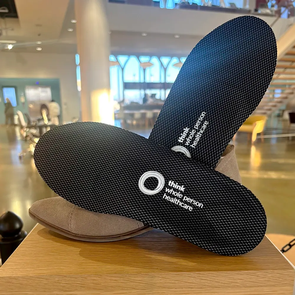 A pair of black insoles with think whole person healthcare printed on them, displayed on top of beige shoes in a brightly lit indoor setting.