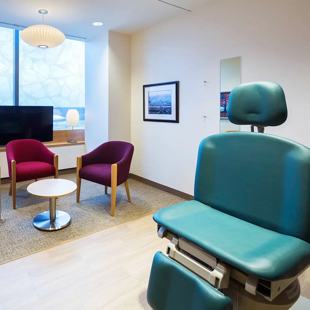 A modern medical exam room with a teal exam chair in the foreground, two purple armchairs and a round table near a window, and soft lighting from a lamp and a ceiling light.