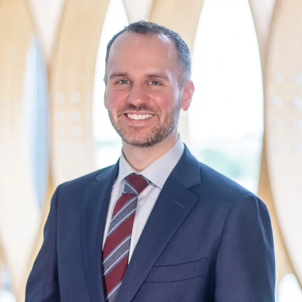 DO Justin Schramm, a man with short brown hair and a beard, stands smiling in a navy suit, striped tie, and light blue shirt before a bright, modern backdrop with curved wooden panels and large windows.