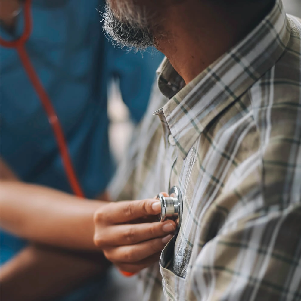 A healthcare professional uses a stethoscope to listen to the chest of an older man wearing a plaid shirt during a medical examination.