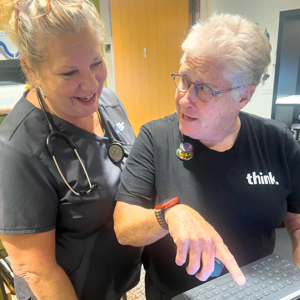 Two older women stand together, smiling and talking. One wears scrubs with a stethoscope and glasses on her head. The other, in a think. shirt, points at a laptop keyboard while looking at her companion.