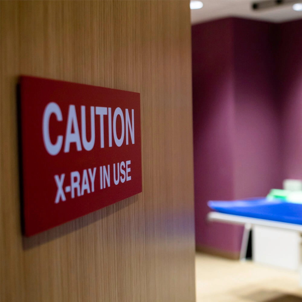 A red sign on a wooden wall reads CAUTION X-RAY IN USE. In the background, there is an examination table in a room with purple walls, indicating a medical or radiology setting.