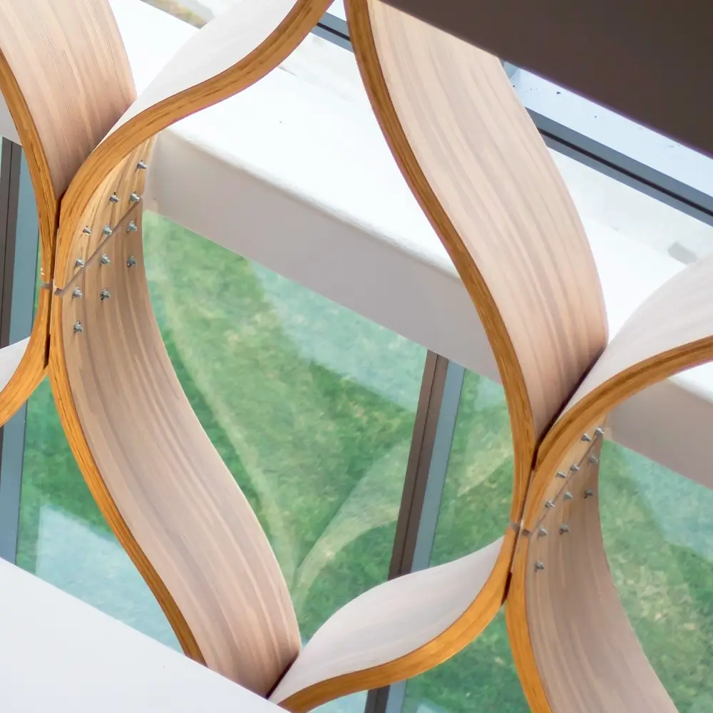 Curved wooden panels with metal bolts form a wavy decorative structure beside large glass windows, through which green grass is visible outside.