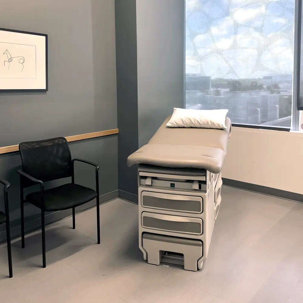 A clean, modern medical exam room with a padded exam table, pillow, two black chairs, a framed line drawing on the wall, and sunlight streaming through a window.