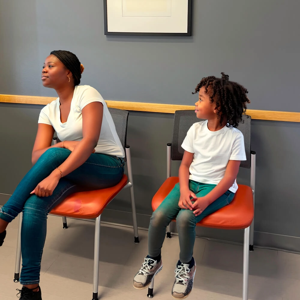 Two people, an adult and a child, sit on orange chairs in a waiting room. Both wear white shirts and jeans. The adult looks to the left, while the child looks at the adult. The wall behind them is gray with a framed picture.