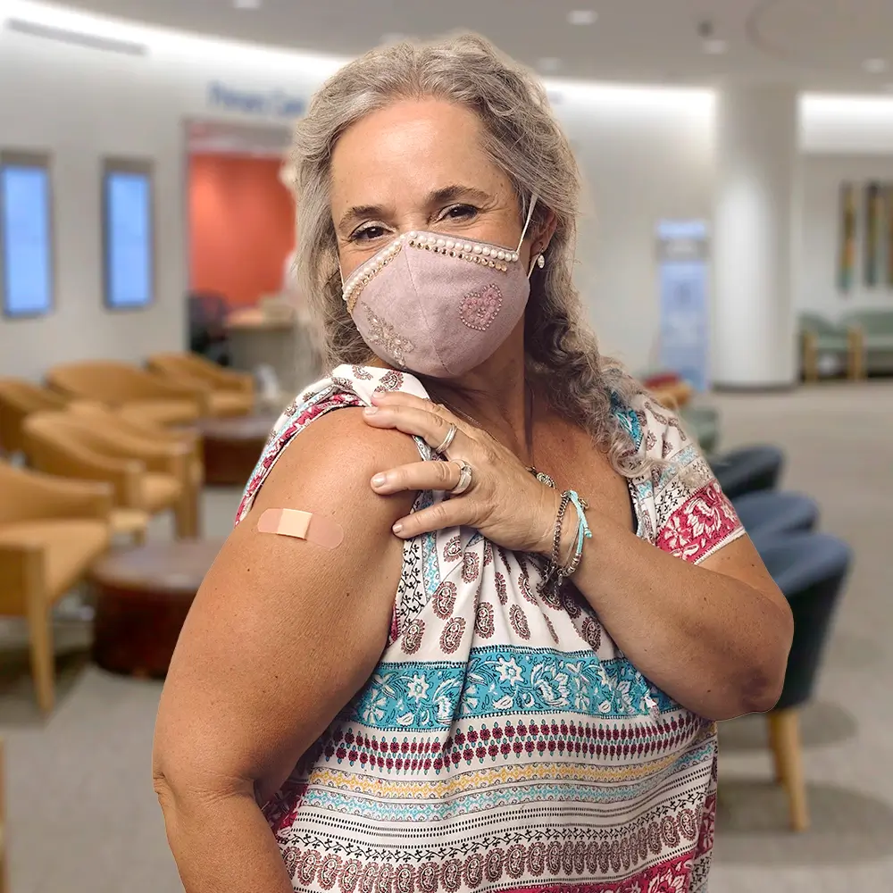 A woman wearing a face mask shows her upper arm with a small bandage, indicating she has received a vaccination. She stands in a waiting area with chairs and soft lighting in the background.