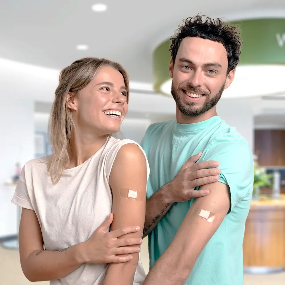 A smiling woman and man stand side by side indoors, each showing a small adhesive bandage on their upper arm, suggesting they have just received vaccinations.