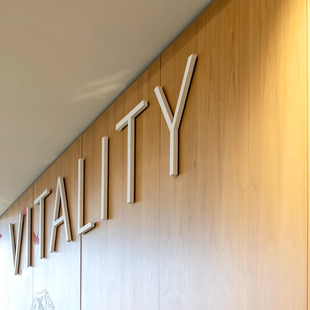 Large white letters spelling VITALITY are mounted on a light wooden wall, viewed at an angle from below. The setting appears to be modern and well-lit.