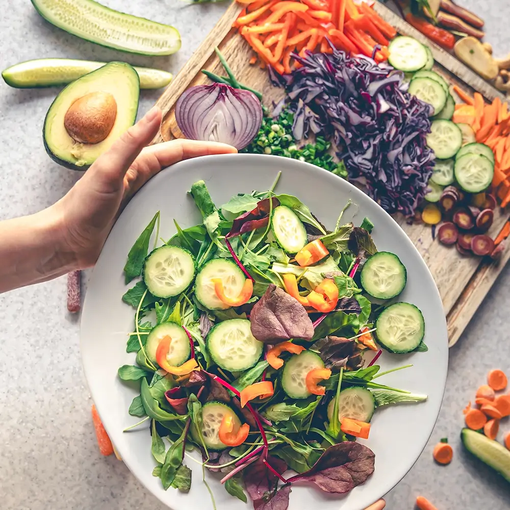 A hand holds a bowl of fresh salad with leafy greens, cucumber slices, and bell peppers. In the background, a cutting board displays chopped vegetables including red cabbage, carrots, onion, and avocado.