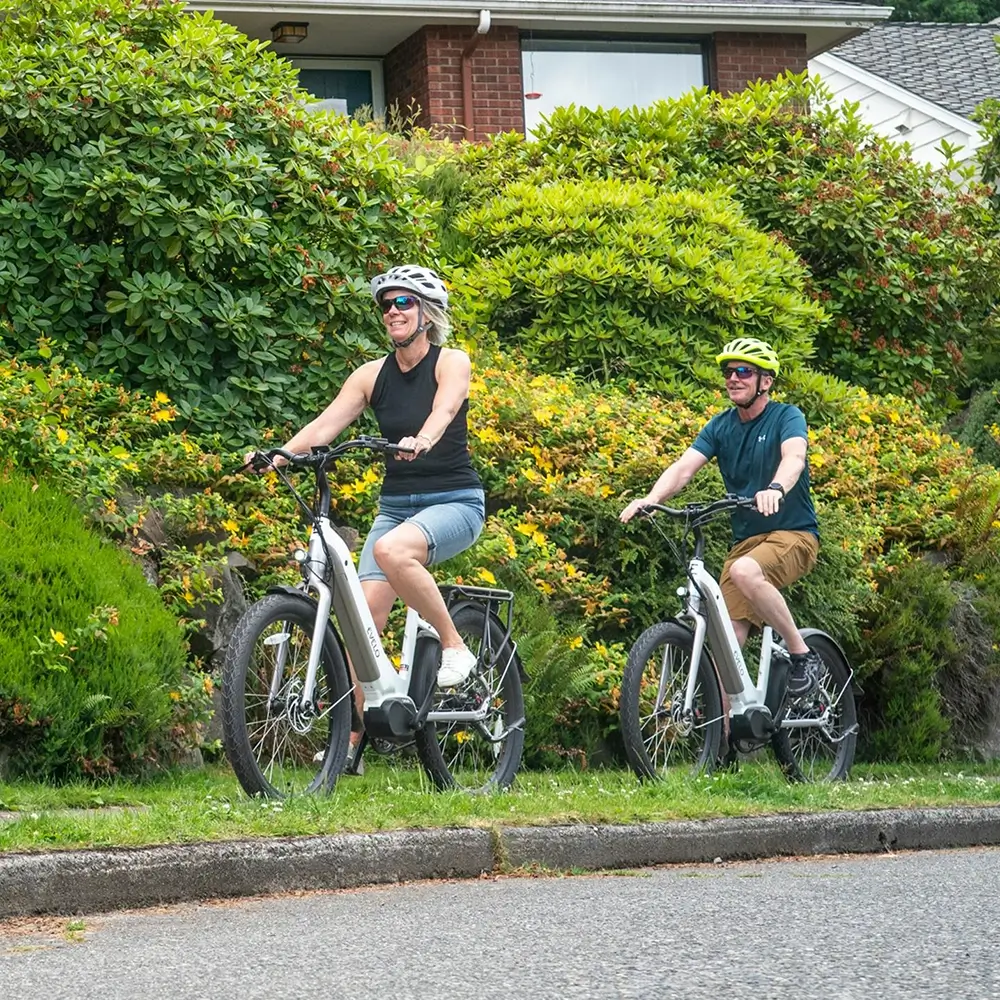 A couple rides electric bikes on a residential street with lush green foliage and yellow flowers in the background.