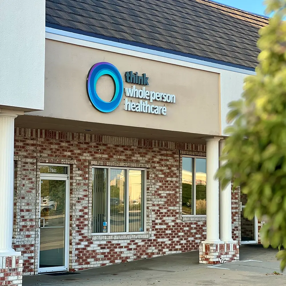 A brick and stucco building with a sign that reads think healthcare above the entrance. A glass door and windows are visible, with part of a leafy tree on the right side.