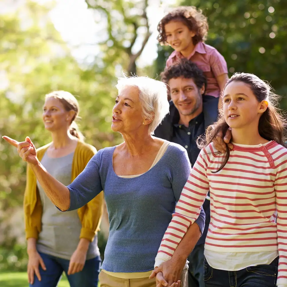 A multigenerational family walks outdoors in a park. An older woman points ahead, a younger girl holds her hand, an adult man carries a child on his shoulders, and another woman walks beside them. All appear happy and engaged.