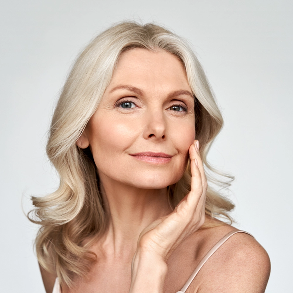 A confident older woman with long, wavy gray hair smiles gently, touching her face with one hand. She wears a light tank top and poses against a plain, light background.