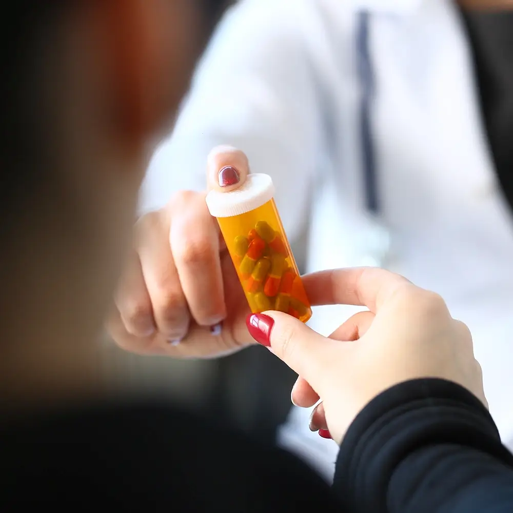 A person hands a prescription pill bottle filled with capsules to another person. A doctor with a stethoscope is visible in the background, slightly out of focus.