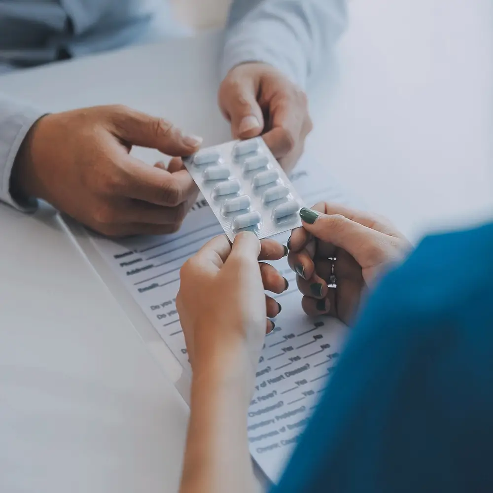 Two people exchange a blister pack of pills over a document on a desk, one wearing a blue sleeve and the other a light-colored sleeve.