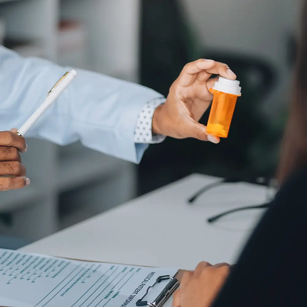 A doctor’s hand holding a prescription pill bottle while talking to a patient, with a clipboard and pen visible on the desk.