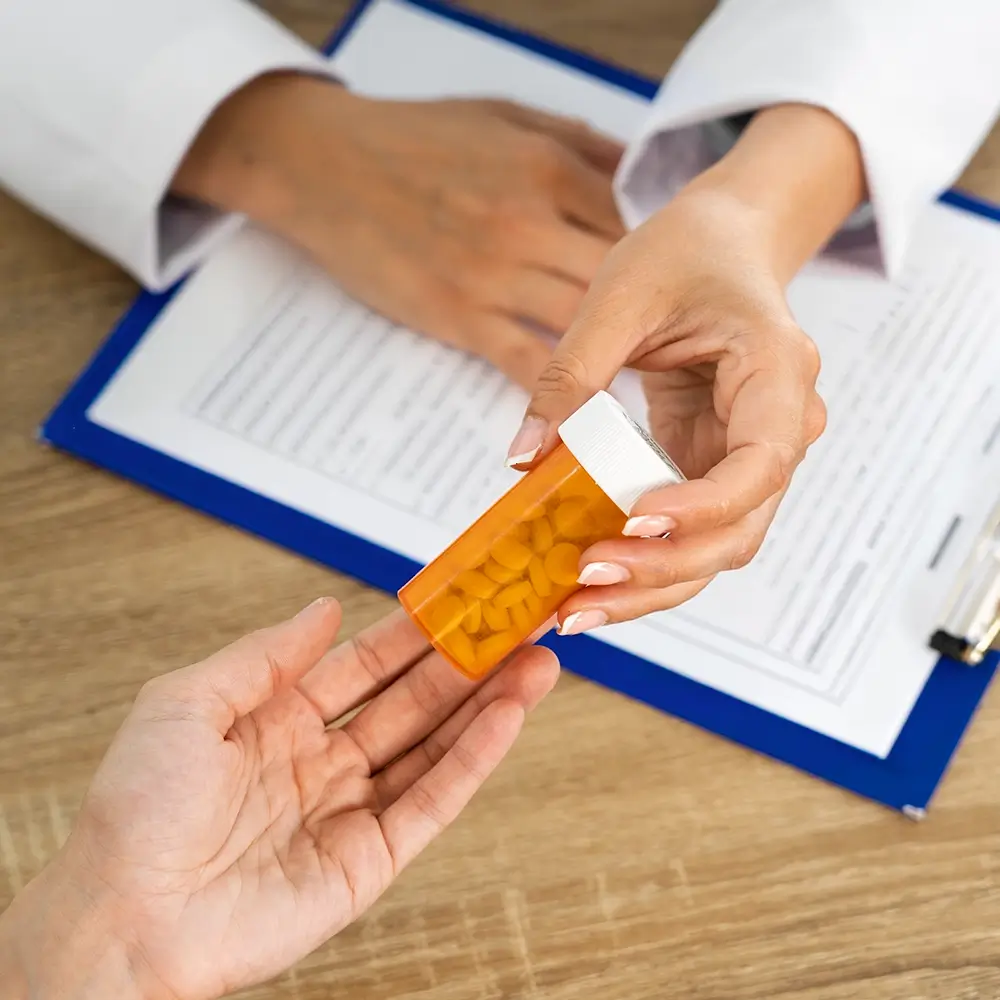 A person hands a bottle of yellow pills to another person across a desk with a clipboard and document.