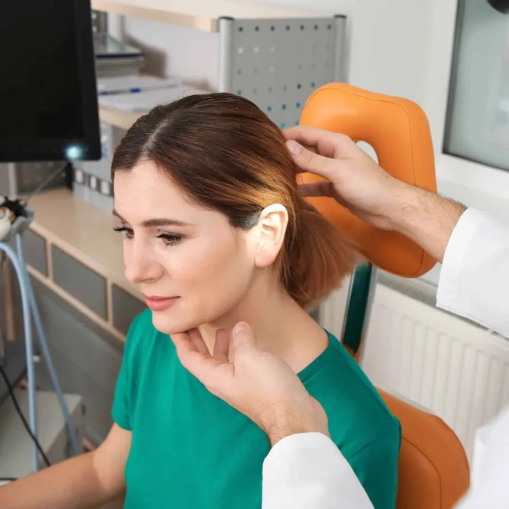 A doctor examines a woman’s ear in a medical office. The woman, wearing a green shirt, sits in an orange chair while the doctor gently touches her ear and chin. Medical equipment is visible in the background.