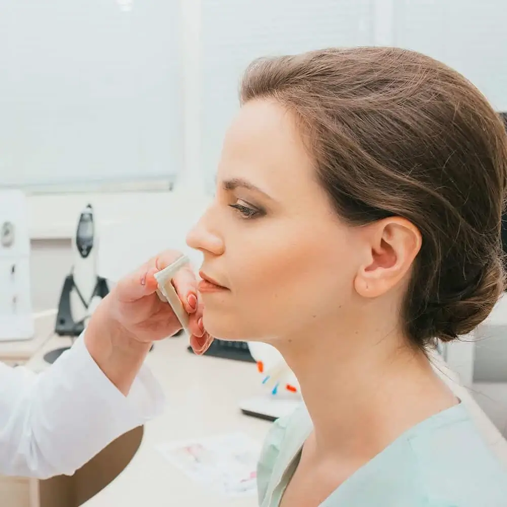 A woman sits in a clinic while a medical professional examines her face with a tool. The setting appears clean and clinical, with medical equipment in the background.