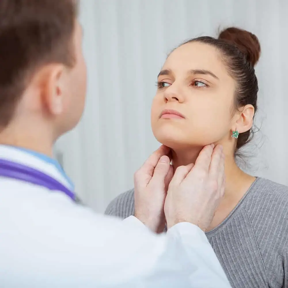 A doctor examines a womans neck, checking for swelling or lumps. The woman looks slightly upward while the doctor gently presses on her neck with both hands.
