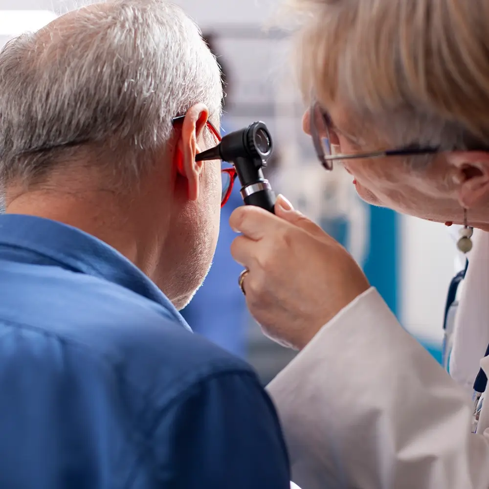 A healthcare professional examines the ear of an older man using an otoscope in a medical setting. The man is seated and facing away from the camera, while the professional focuses closely on his ear.