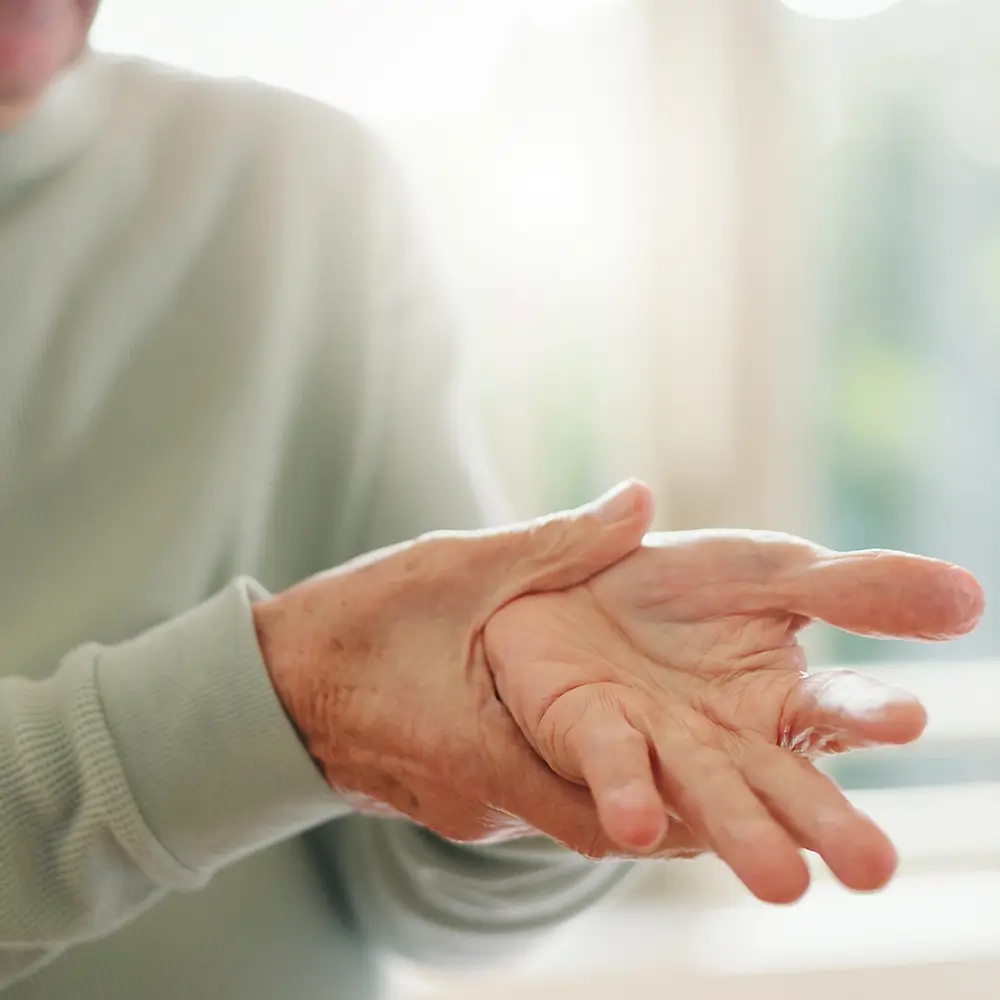 An elderly person holding their left hand with their right hand, appearing to massage or soothe discomfort, with a soft focus background and natural light.