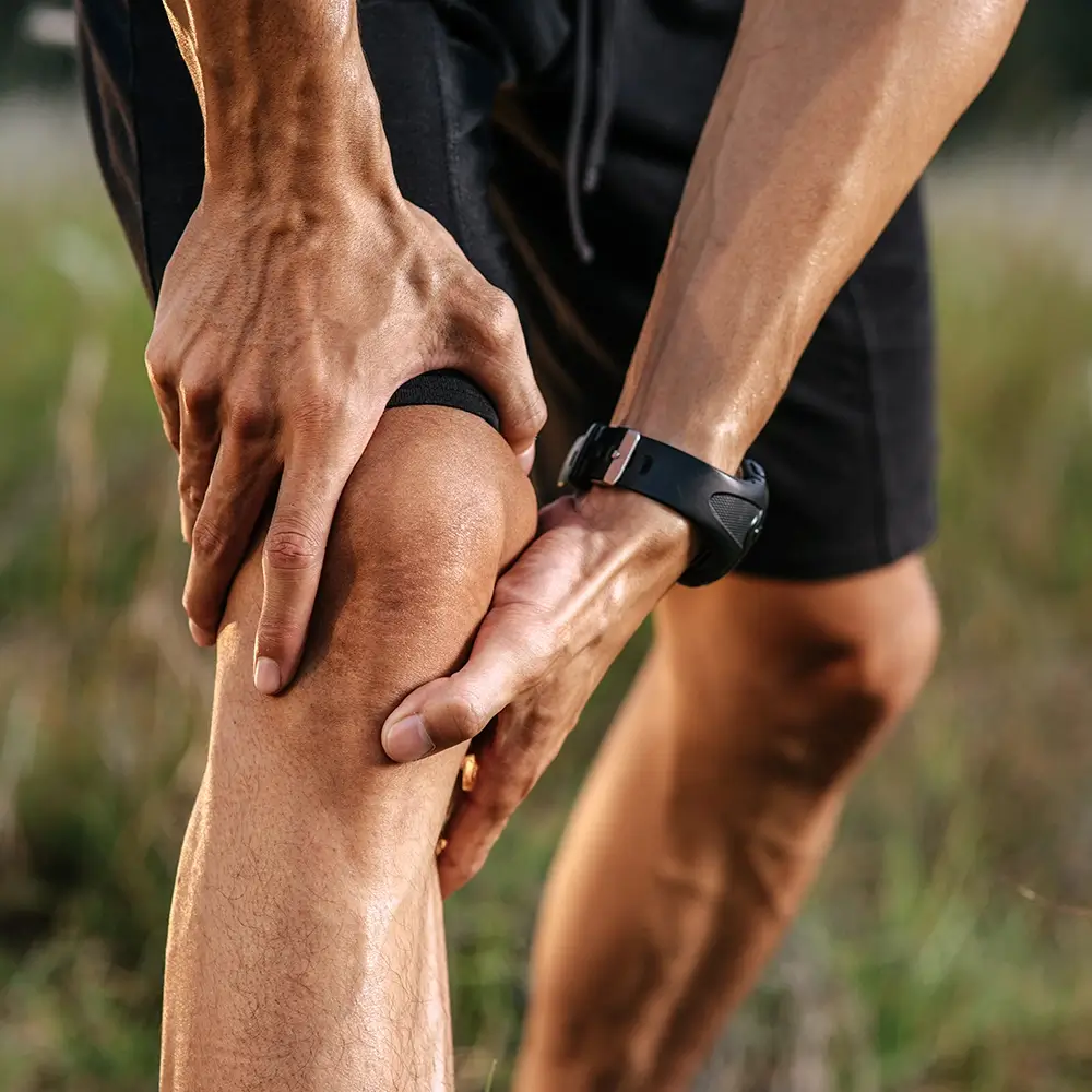 A person wearing black shorts and a wristwatch holds their knee and lower leg, possibly indicating leg pain or injury, outdoors with a blurred background.