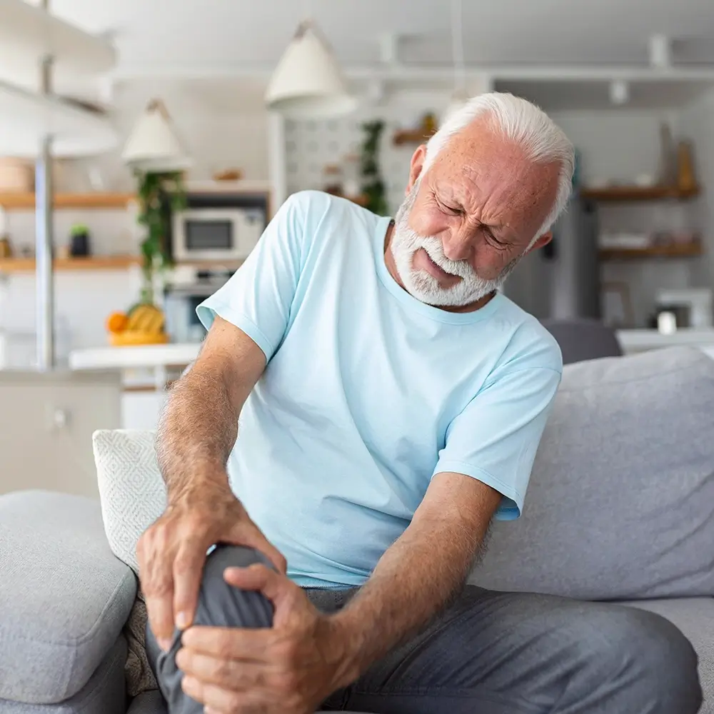 An elderly man with white hair and a beard sits on a couch, grimacing in pain while holding his knee with both hands, suggesting discomfort or injury.