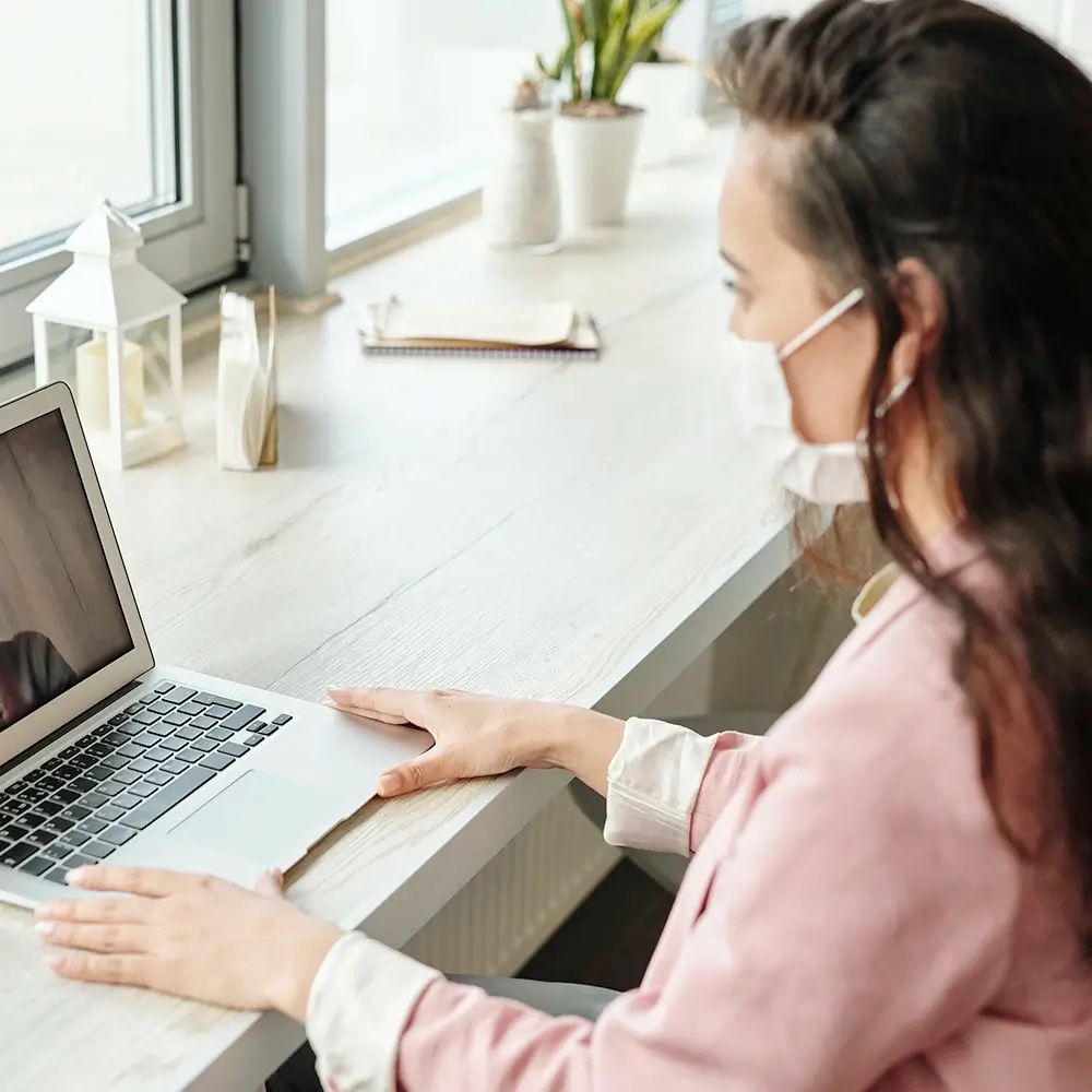 A woman wearing a face mask sits at a desk by a window, using a laptop for a video call. She is dressed in a pink top and has long dark hair. The desk has decor items and a notebook on it.