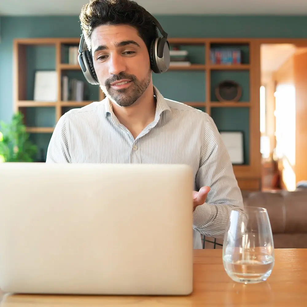 A man wearing headphones sits at a table in front of a laptop, gesturing with his hand, with a glass of water nearby. He appears to be on a video call in a modern, sunlit room with shelves in the background.