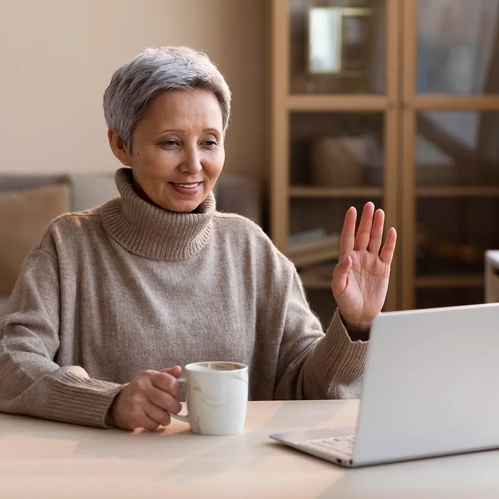 An older woman with short gray hair, wearing a beige turtleneck sweater, sits at a table holding a mug and smiling while waving at a laptop screen during a video call.
