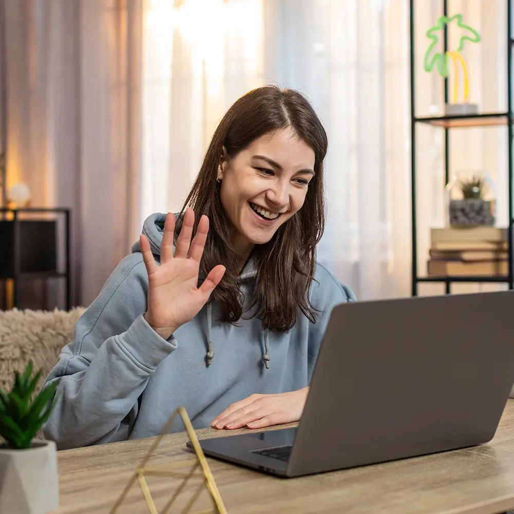 A smiling woman wearing a hoodie sits at a desk, waving at her laptop during a video call. The room has soft lighting, plants, and shelves in the background.