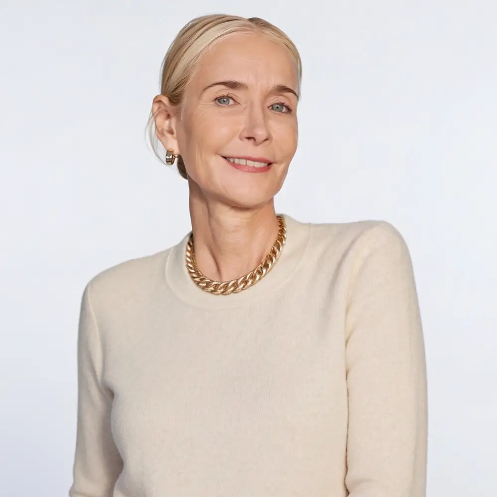 A smiling woman with blonde hair pulled back, wearing a cream sweater, a chunky gold necklace, and gold earrings, stands against a plain light background.