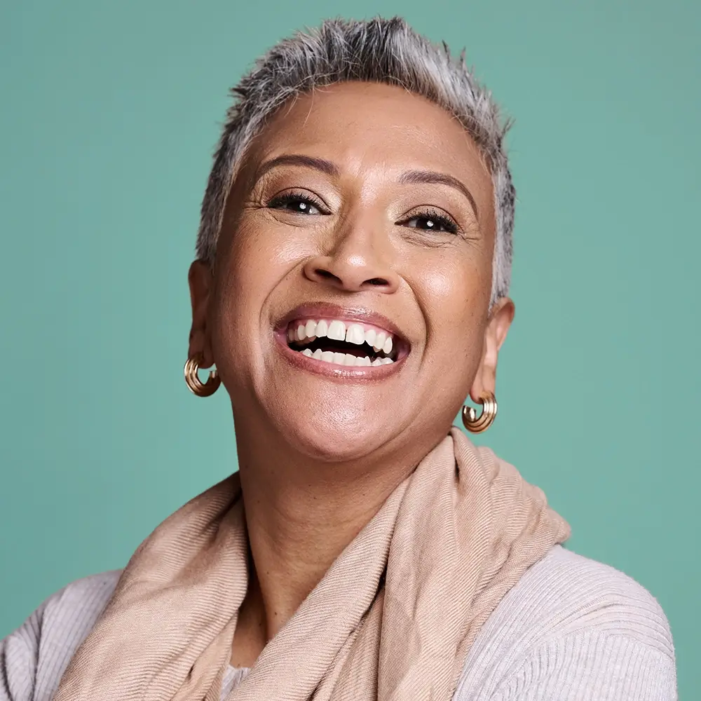 A smiling person with short gray hair, wearing gold hoop earrings, a beige scarf, and a light sweater, poses in front of a plain turquoise background.