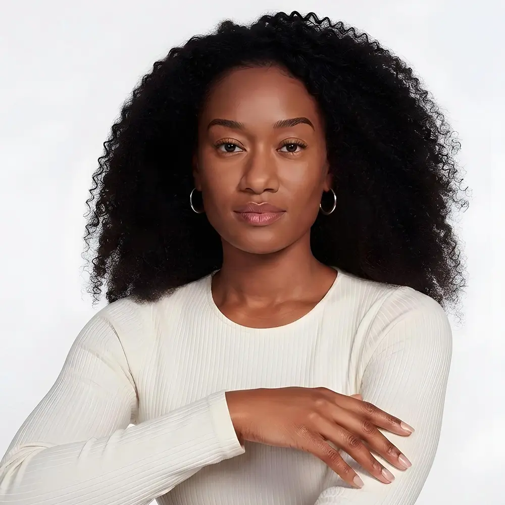 A woman with curly black hair wearing hoop earrings and a long-sleeve, off-white ribbed shirt, poses with her arms crossed in front of a plain, light background.
