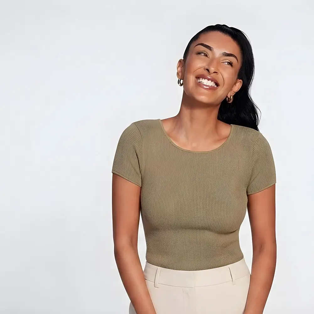 Smiling woman with long dark hair, wearing gold hoop earrings, a fitted short-sleeve olive green top, and high-waisted beige pants, stands against a plain light background.