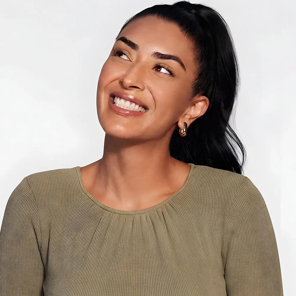 A woman with long black hair in a ponytail, wearing a light olive-green ribbed top and gold hoop earrings, smiles and looks upward against a plain light background.