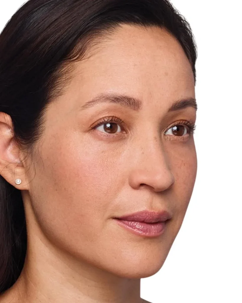 A woman with straight dark hair, clear skin, and brown eyes looks slightly to the side. She has minimal makeup and wears a small stud earring. The background is white.